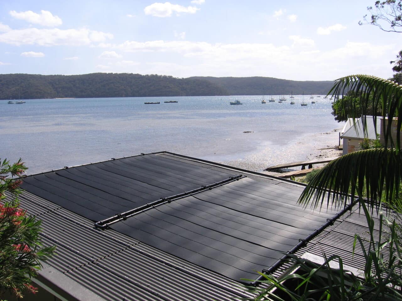 A view overlooking a tranquil lake from behind a dark gray metal rooftop equipped with solar panels, featuring boats on water and a lush green forest in the background under a cloudy sky. | AES - Pool Heating & Energy Efficient Products