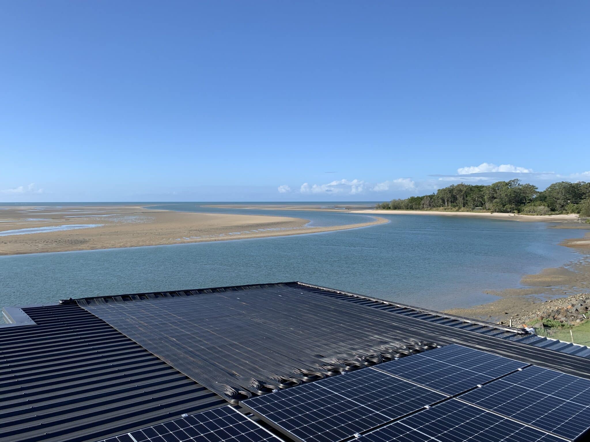 Aerial view of Ultra Solar panels on a rooftop overlooking a serene coastal landscape with a wide, meandering river and tidal flats under a clear blue sky. | AES - Pool Heating & Energy Efficient Products
